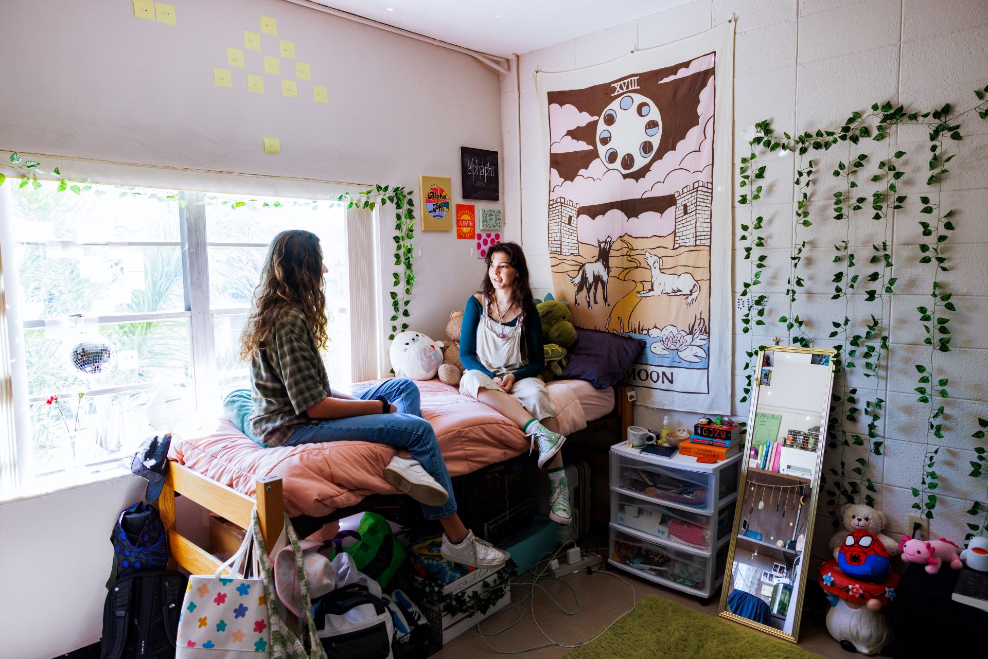 Two young women sitting on a bed in a brightly decorated dorm room, talking and surrounded by plush toys, posters, and hanging plants